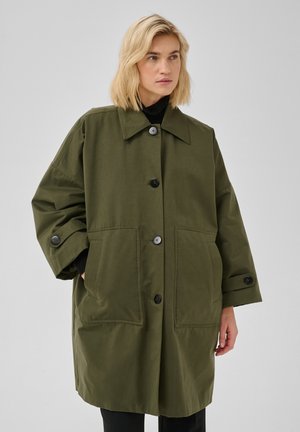 Femme blonde portant un manteau ample vert olive boutonné avec de grandes poches avant et des manches noires visibles en dessous.