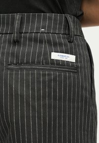 Τσέπη σε παντελόνι με μαύρες ρίγες, ετικέτα με λευκό χρώμα "ELEMENT CO. SINCE 1992" και θηλιές για ζώνη σε άτομο που φοράει σκούρο πάνω μέρος.