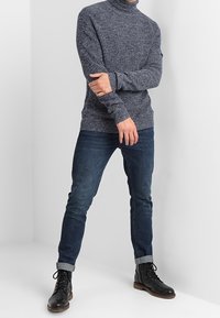 Granatowy sweter z golfem o dzianinowym wzorze, w połączeniu z ciemnoniebieskimi dżinsami i czarnymi butami na sznurówki. Casulowy styl z podwiniętymi mankietami.