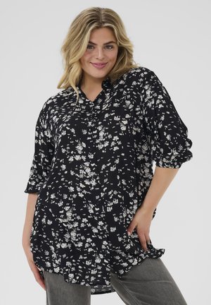 Chemise noire à manches courtes avec un motif floral blanc, manches retroussées, coupe décontractée et ourlet arrondi. Le tissu semble lisse et léger.