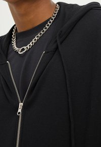 Svart zip-up hoodie med en jämn textur, metalldragkedja och huva. Bärs över en svart skjorta, ihop med ett kraftigt silverhalsband.