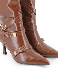 Las botas ankle de imitación cuero marrón presentan una punta afilada y un tacón delgado, con tres detalles de correas ajustables acentuados por herrajes de color dorado.