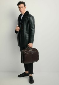 Czarny, dwurzędowy płaszcz ze skóry z kołnierzem ze shearlingu, trzymający brązową skórzaną teczkę. Czarnie spodnie i ciemne sneakersy dopełniają stylizację.