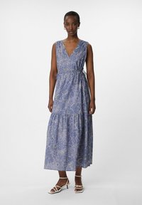 Vestito blu a fantasia floreale con scollatura a V, cintura in vita e gonna a balze. Realizzato in tessuto leggero. Indossato con tacchi bianchi con cinturini.