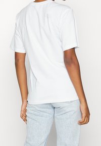 Bílý tričko s krátkým rukávem a kulatým výstřihem. Na horní části zad je vytištěno malé logo v světle šedé barvě. Vyrobeno z bavlněného materiálu.