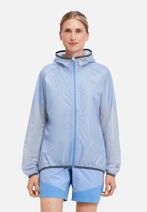 Frau mit blonden Haaren, die eine hellblaue, mit Kapuze versehene Windjacke und passende Shorts trägt, vor einem schlichten weißen Hintergrund stehend.