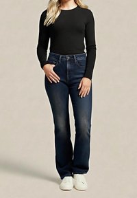 Femme portant un haut à manches longues noir, un jean bleu foncé et des baskets blanches, debout avec une main dans la poche, sur un fond uni.