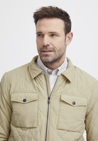 Beige tikandatud jakk, millel on krae, kaks klapiga taskut ja eesne åpärukett. Seljas triibuline särk, millel on tekstuuriga pind.