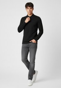 Czarna polo z długim rękawem i kołnierzykiem, z subtelnym logo. W zestawie z ciemnoszarymi wąskimi dżinsami i białymi sneakersami na neutralnym tle.