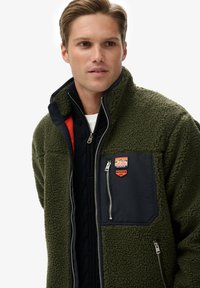 Giacca in pile verde con tasca sul petto blu navy, davanti con cerniera e logo arancione. Tessuto strutturato, collo alto e design a strati.