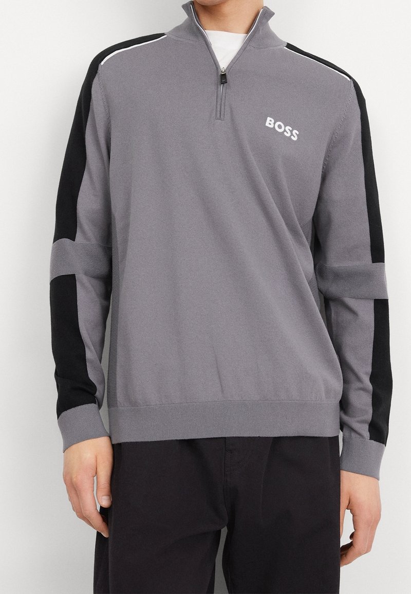 Mann in grauem und schwarzem Langarm-Pullover mit Viertelreißverschluss und "BOSS"-Logo auf der linken Brust, kombiniert mit schwarzen Hosen.
