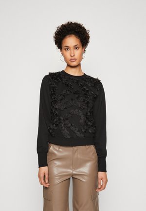 Black sweatshirt with textured, ruffled floral accents on the front, long sleeves, and ribbed cuffs. Paired with smooth beige, high-waisted pants.
