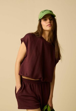 Femme portant un sweat sans manches bordeaux et un short assorti, casquette verte avec le texte « AMOR », tenant un sac vert sur un fond beige uni.