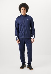 Chándal atlético de color azul marino, hecho de una tela suave y ligera. Presenta una chaqueta con cremallera y detalles en blanco, junto con pantalones a juego. Zapatillas negras.