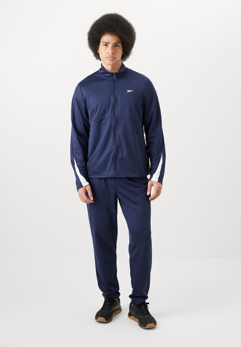 Chándal atlético de color azul marino, hecho de una tela suave y ligera. Presenta una chaqueta con cremallera y detalles en blanco, junto con pantalones a juego. Zapatillas negras.