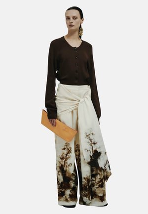 Frau trägt ein dunkelbraunes, geknöpftes Oberteil, weite cremefarbene Hose mit braunem Landschaftsprint und langem Bindeband, hält eine beige Clutch und große goldene Ohrringe.