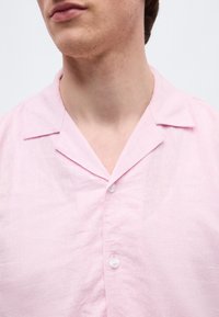 Κοντινό πλάνο ενός ατόμου που φοράει ροζ ανοιχτό πουκάμισο με γιακά, κουμπωμένο με δύο λευκά κουμπιά, εμφανίζοντας το λαιμό και το κάτω μέρος του προσώπου.