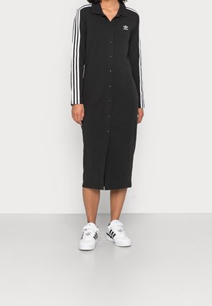 Žena v černých adidasových šatech s dlouhým rukávem na knoflíky s bílými pruhy na rukávech a bílými teniskami Adidas s černými pruhy.