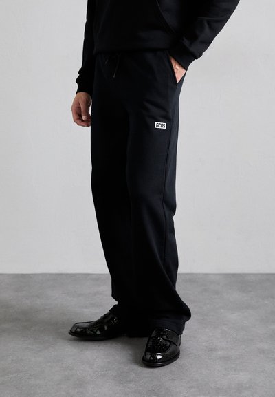 Pantaloni tuta neri con vita regolabile, caratterizzati da un piccolo logo "GCOS" sull'anca. Tessuto liscio e design a gamba dritta. Scarpe nere con finitura lucida.