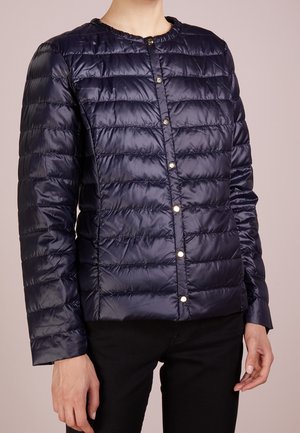 Kvinde iført en marineblå, langærmet, quiltet jakke med trykknapper, kombineret med sorte bukser, stående mod en ensfarvet baggrund.