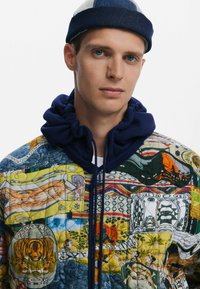 Giacca a cappuccio trapuntata con un design patchwork colorato, caratterizzata da varie fantasie, prevalentemente nei toni del giallo, blu e verde, con accenti blu navy.