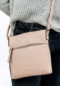Borsa a tracolla in pelle rosa chiaro con tasca frontale con cerniera, design piatto e hardware in argento. Indossata con un maglione grigio in maglia e jeans scuri.