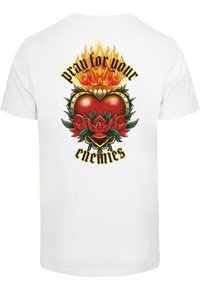 Camiseta blanca con un gráfico en la parte posterior de un corazón rojo con llamas, rodeado de hojas verdes y rosas rojas, con el texto "reza por tus enemigos."