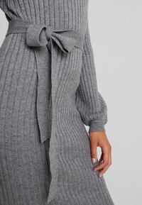 Robe en maille côtelée grise avec une taille nouée, des manches longues et une texture douce. Le tissu présente des rayures verticales pour plus de détail.