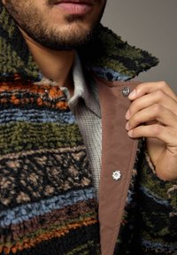 Getextureerde, meerkleurige jas met strepen van zwart, groen, blauw en oranje. Heeft een bruine binnenvoering en een drukknoopsluiting bij de kraag.
