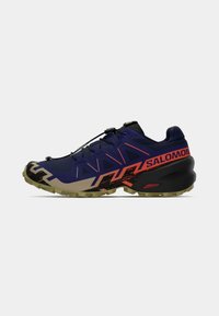 Sapatilha de trail running em roxo e preto com toques laranja, sola de borracha texturizada, sistema de atilhos seguro e um design anguloso distinto.