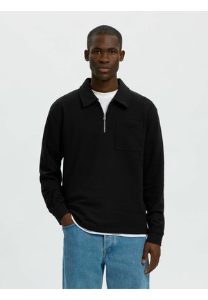 Homme portant une chemise noire zippée à manches longues avec poche poitrine par-dessus un tee-shirt blanc et un jean bleu, debout avec une main dans la poche sur un fond uni.