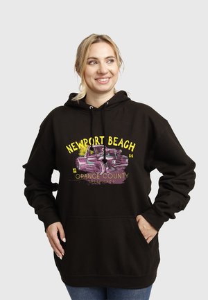 Donna sorridente che indossa una felpa nera con cappuccio con la scritta "Newport Beach Orange County" e un design di un'auto d'epoca viola, in piedi con le mani nelle tasche.