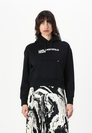 Svart cropped hoodie laget av sweatshirt-stoff med en frontlomme og hvit "KARL LAGERFELD JEANS" logo. Kombinert med et plisserte svart og hvitt skjørt.