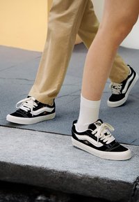 Swarte og kvite sneakers med blomsterbroderi, laget av lerret og semsket skinn, teksturerte svarte tåhette, og kvite snørebånd, kombinert med lyse sokker.