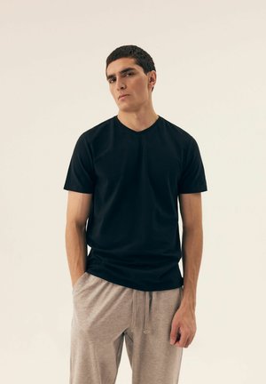 Schwarzes T-Shirt mit kurzen Ärmeln und V-Ausschnitt, aus weichem Stoff gefertigt, getragen mit hellgrauen Jogginghosen mit Kordelzug. Das Model steht mit den Händen in den Taschen.