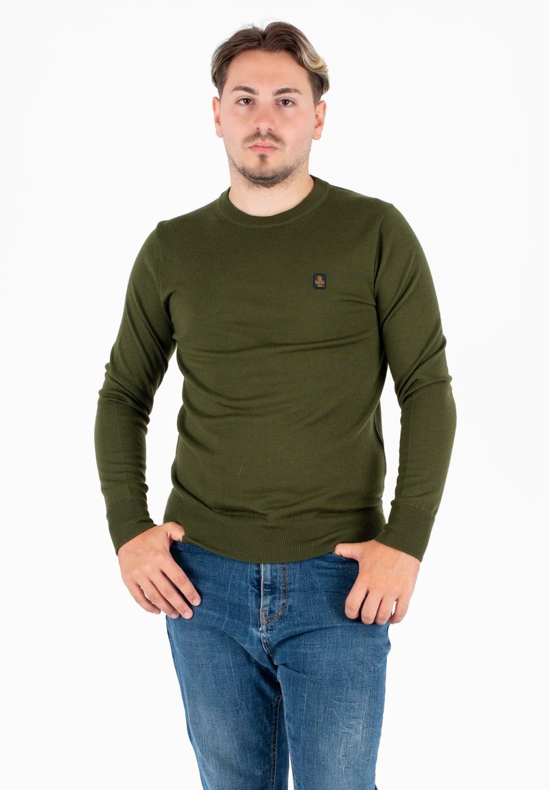 Jonge man met kort haar die een olijfgroene lange mouwen trui en blauwe jeans draagt, staand met handen dicht bij zijn zakken tegen een witte achtergrond.