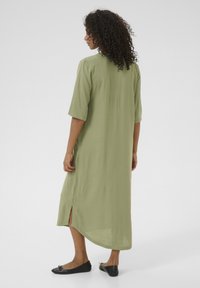 Robe verte olive, longueur genoux, à manches courtes, avec fentes latérales et coupe droite. Portée avec des chaussures plates noires.