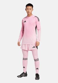 Maglia sportiva rosa a maniche lunghe, con strisce nere e logo; abbinata a pantaloni corti rosa e leggings, accentuata da scarpe da calcetto nere.