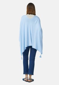 Kobieta ubrana w jasnoniebieski szal z frędzlami, beżową bluzkę, ciemnoniebieskie spodnie typu cropped i niebieskie sandały płaskie, stojąca tyłem.