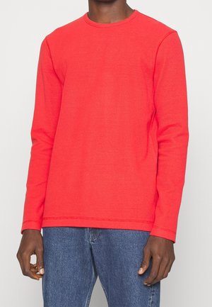 Mand iført en knald rød langærmet skjorte og blå jeans, stående mod en ensfarvet lys baggrund.