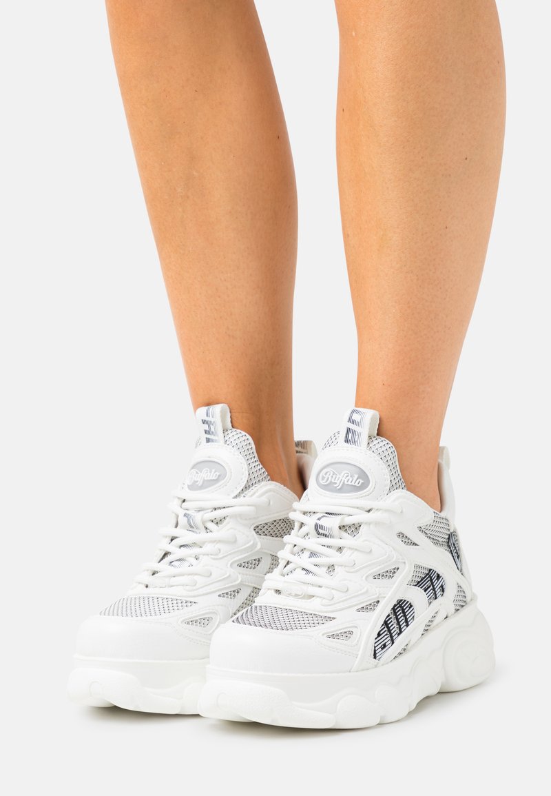 Białe sportowe buty z siateczkowej tkaniny, masywną podeszwą i widocznymi sznurówkami. Zawierają logo na języku i pięcie, z teksturowanymi akcentami.