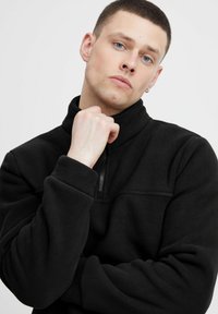 Svart fleece genser med høy krage og halv glidelås foran. Den har lange ermer og en myk tekstur. Minimalistisk design uten mønstre.
