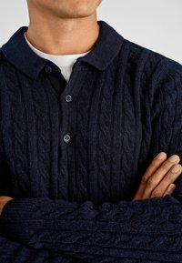 Marineblauer Pullover im Zopfmuster mit Kragen, drei Knöpfen und gerippten Bündchen. Strukturierte Wolle verleiht dem Design Tiefe.