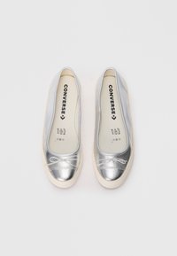 Silberne, metallische Ballerina-Converse-Schuhe mit weißen Gummi-Sohlen und kleinen dekorativen Schleifen auf einem schlichten weißen Hintergrund.