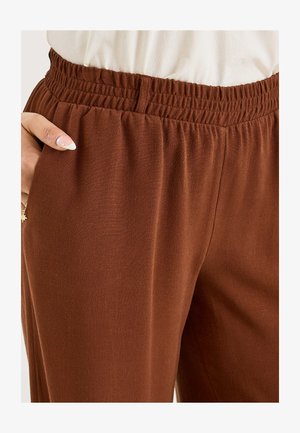 Personne portant un pantalon marron à taille élastique, la main gauche dans la poche, montrant un ongle blanc avec une petite décoration en perle.