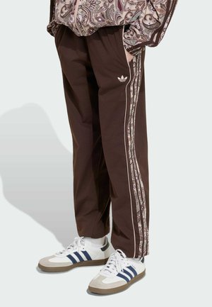 Person iført brune Adidas løbebukser med mønstrede striber på siden og hvide Adidas sneakers med blå striber, stående med hænderne i lommerne.