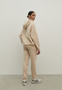 Bežová mikina s kapucňou a voľným strihom, spojená s zodpovedajúcimi bežovými teplákmi. Mäkko spracovaná textúra a pružné manžety. Doplnené športovými topánkami.