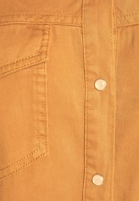 Gros plan sur un tissu orange avec des coutures détaillées, un coin de poche et deux boutons-pression métalliques ronds sur un vêtement.