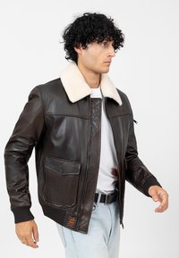 Bruine leren bomberjack met een crème schapenvacht kraag, geribde manchetten, twee voorkruiszakken en een ritsluiting. Gestyled met lichte jeans.