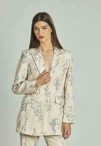 Blazer di colore crema in tessuto leggero, con intricato ricamo floreale blu, rever affilati e due tasche frontali.